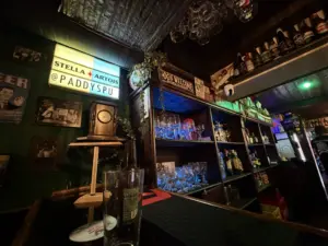 A dimly lit bar with shelves stocked with bottles and glasses, illuminated blue lights, a clock, vintage signs, and a glowing “Stella Artois @PADDYSPU” sign above the bar counter.