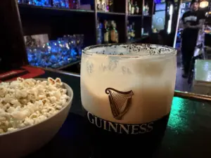 A close-up of a pint of Guinness stout beer with a creamy head, next to a bowl of popcorn on a bar counter; shelves with bottles and glasses are visible in the background.